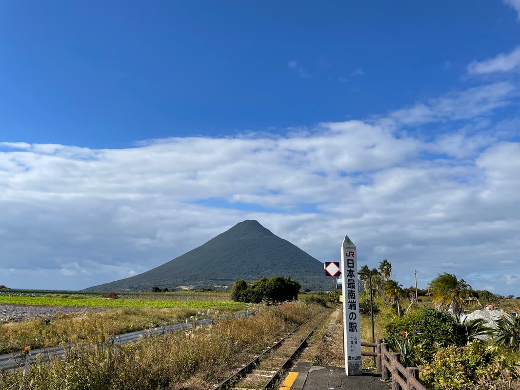 This is a photo showing the small station building of JR Nishi-Ōyama Station with surrounding rice fields and Sakurajima volcano in the background.